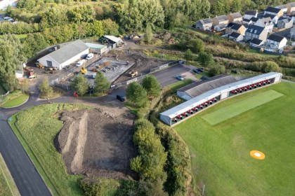 Aerial view of the Machynys golf club driving range, with a fenced building site and concrete slab beside the club's maintenance shed, and houses beyond