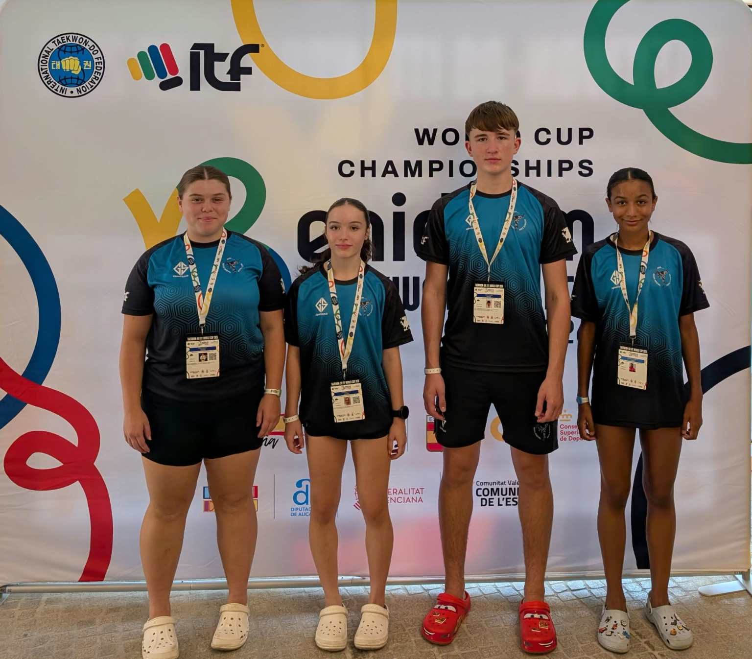 Four young taekwon-do athletes in matching teal and black club shirts stand in front of the ITF Taekwon-Do World Cup board in Benidorm