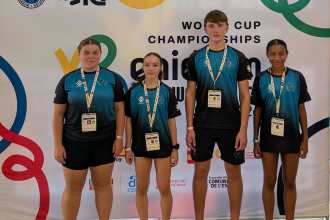 Four young taekwon-do athletes in matching teal and black club shirts stand in front of the ITF Taekwon-Do World Cup board in Benidorm