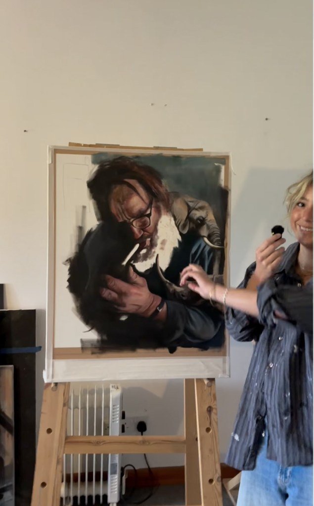 A smiling young woman points to an unfinished painting on an easel of a bearded man in glasses with a small elephant