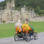 A family rides a four-wheeled pedal cart along the path in front of Margam Castle