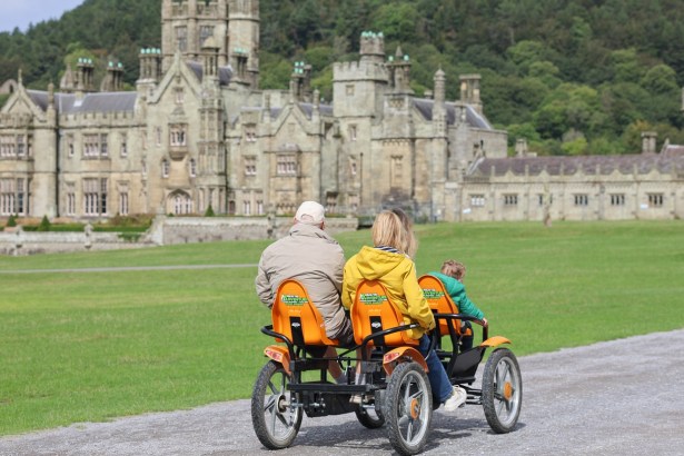 A family rides a four-wheeled pedal cart along the path in front of Margam Castle