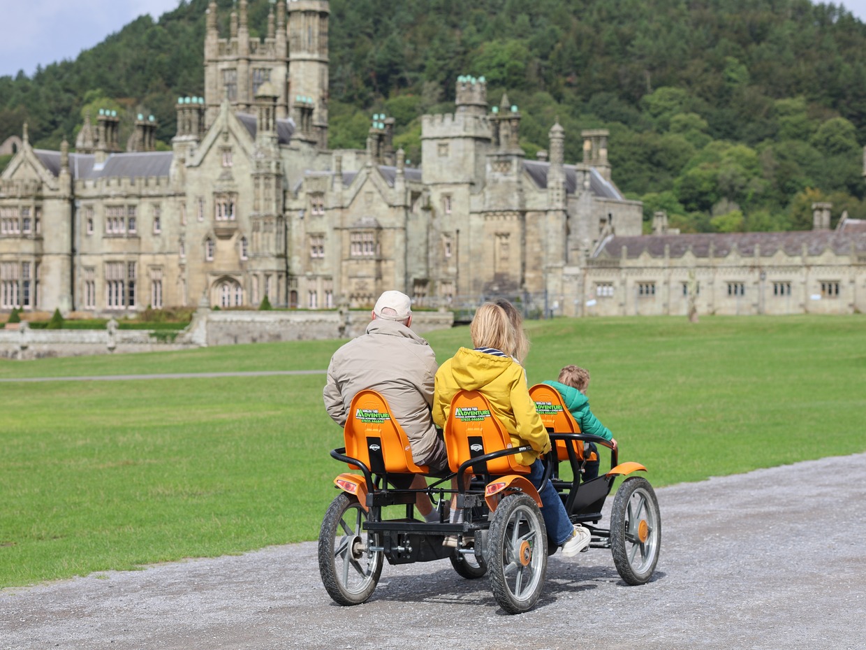 A family rides a four-wheeled pedal cart along the path in front of Margam Castle