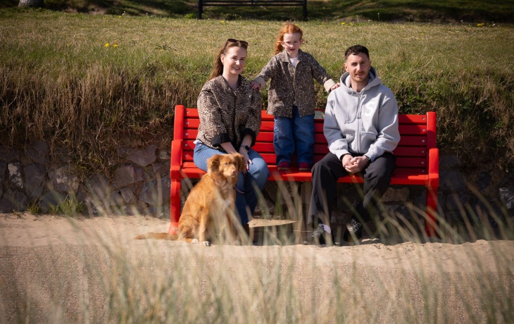 Emily and Mathew sitting on a red bench on the sand at Swansea Beach, with Esme standing between them and their dog sitting in front