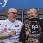 Mark Francis in a white Ospreys shirt and Helen Francis in an Ospreys scarf laughing in stadium seats, each holding a glass of champagne