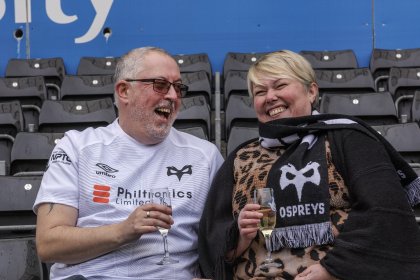 Mark Francis in a white Ospreys shirt and Helen Francis in an Ospreys scarf laughing in stadium seats, each holding a glass of champagne