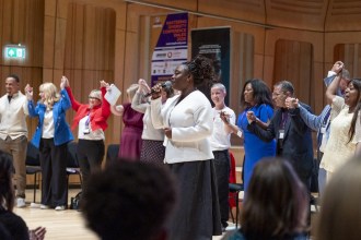 Maryann Enereba, in a white jacket, sings into a microphone while a line of delegates behind her hold hands raised in the air in a wood-panelled hall