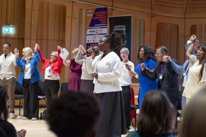 Maryann Enereba, in a white jacket, sings into a microphone while a line of delegates behind her hold hands raised in the air in a wood-panelled hall
