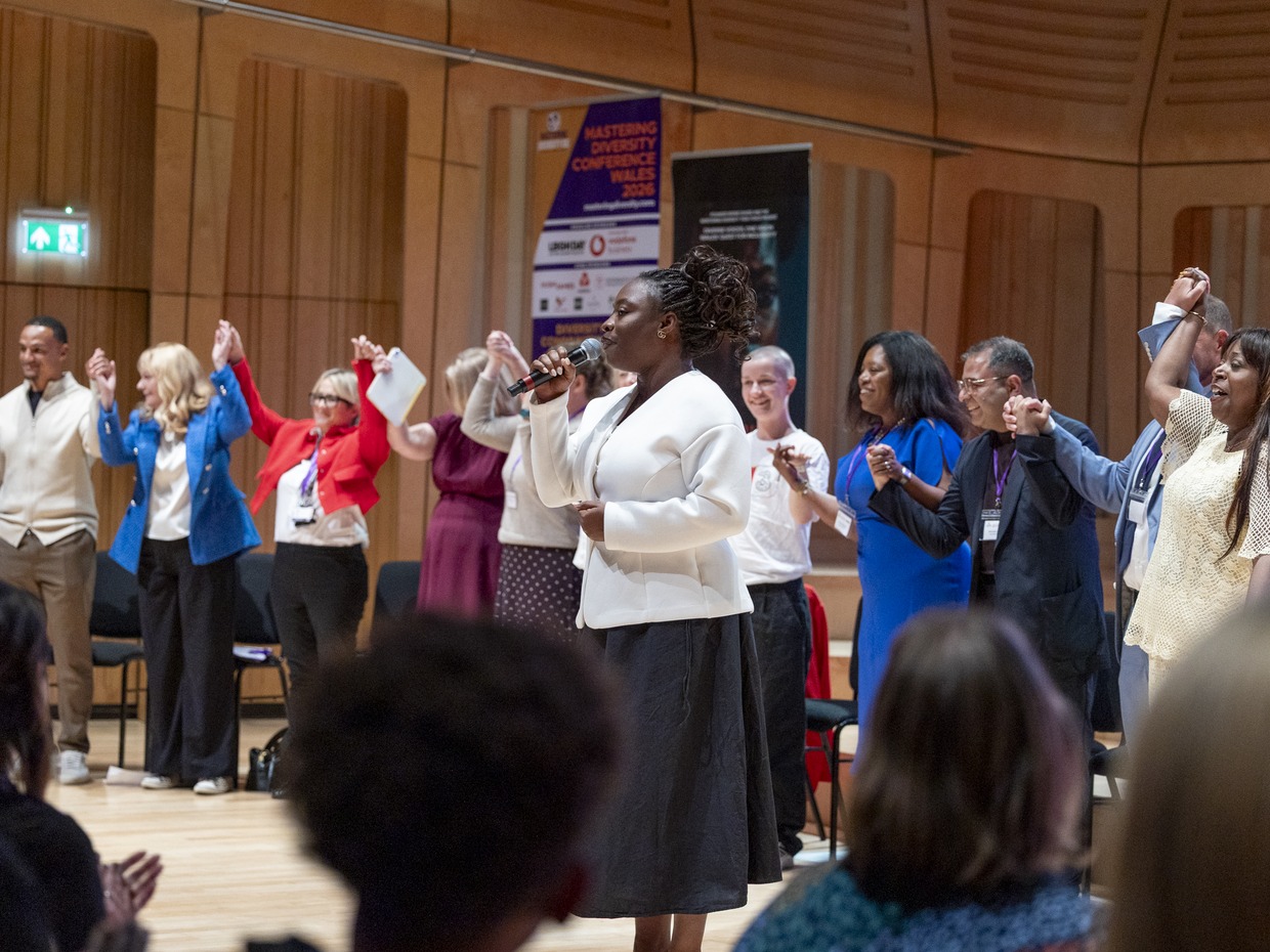 Maryann Enereba, in a white jacket, sings into a microphone while a line of delegates behind her hold hands raised in the air in a wood-panelled hall