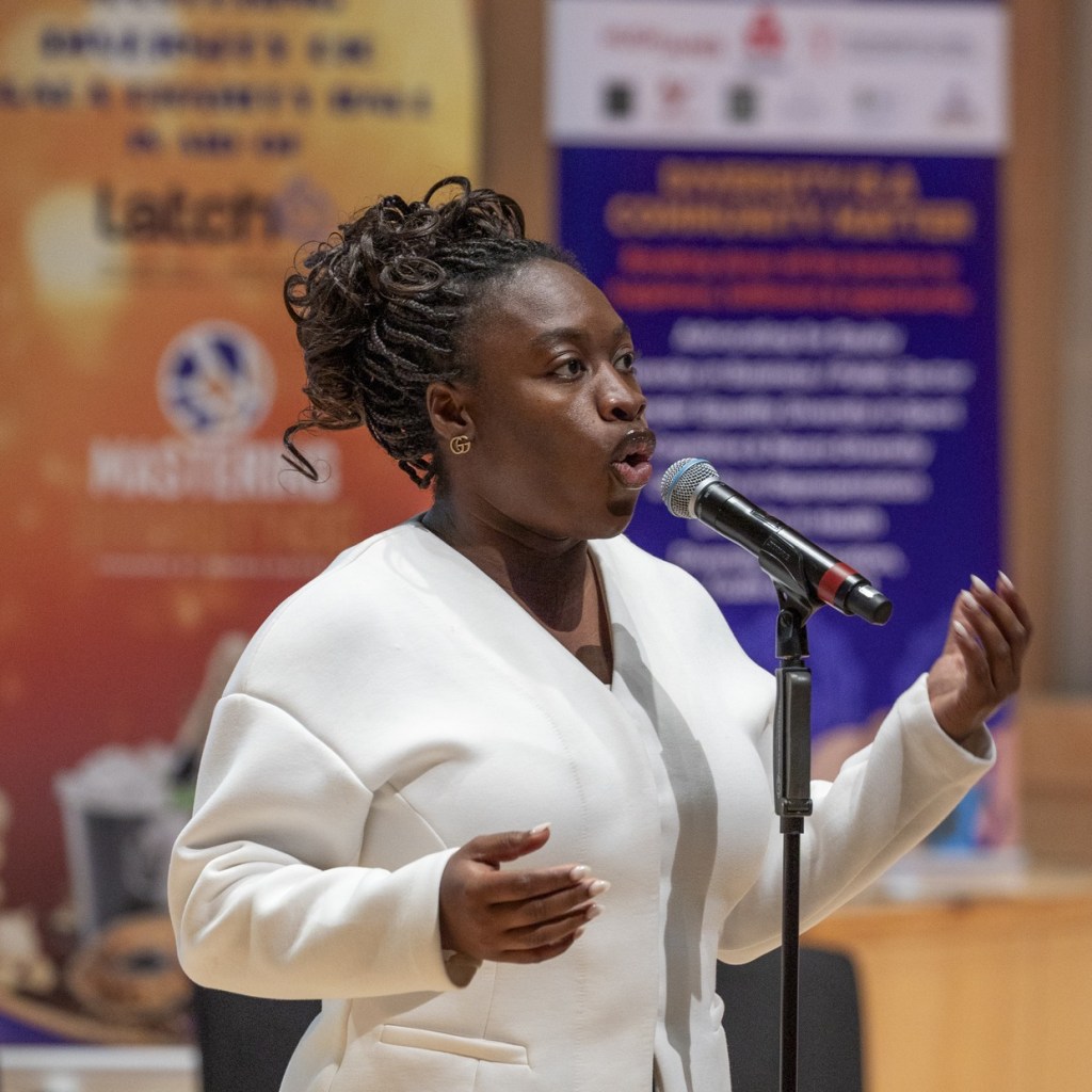 Maryann Enereba in a white jacket singing into a microphone on a stand, with event banners behind her