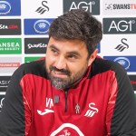 Swansea City head coach Vitor Matos, in a red and black training top, smiles at a press conference in front of a sponsor board