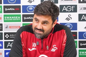 Swansea City head coach Vitor Matos, in a red and black training top, smiles at a press conference in front of a sponsor board