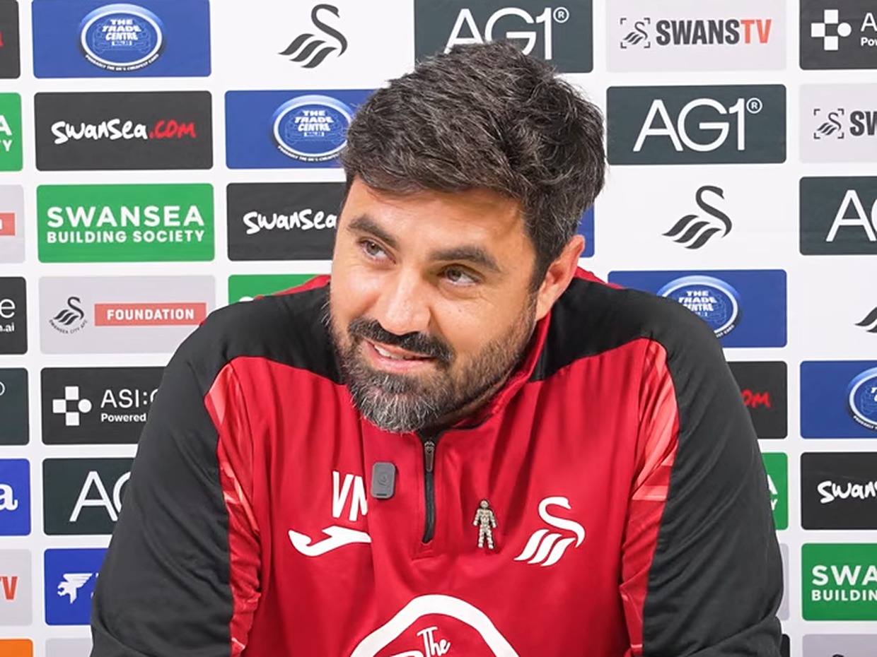 Swansea City head coach Vitor Matos, in a red and black training top, smiles at a press conference in front of a sponsor board