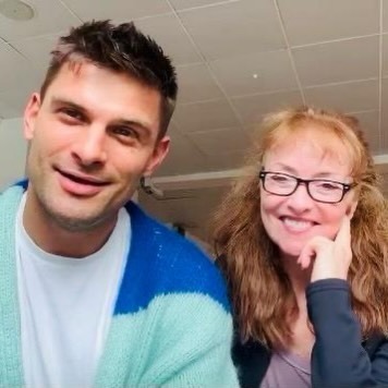 A man in a blue and mint cardigan and a smiling woman in glasses pose together for a selfie in a rehearsal room