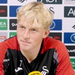 Swansea City's Melker Widell at a press conference in front of a sponsor board, wearing a red and black training top