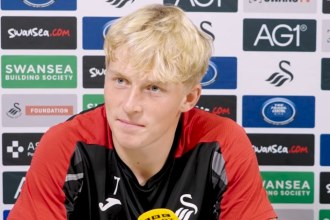 Swansea City's Melker Widell at a press conference in front of a sponsor board, wearing a red and black training top