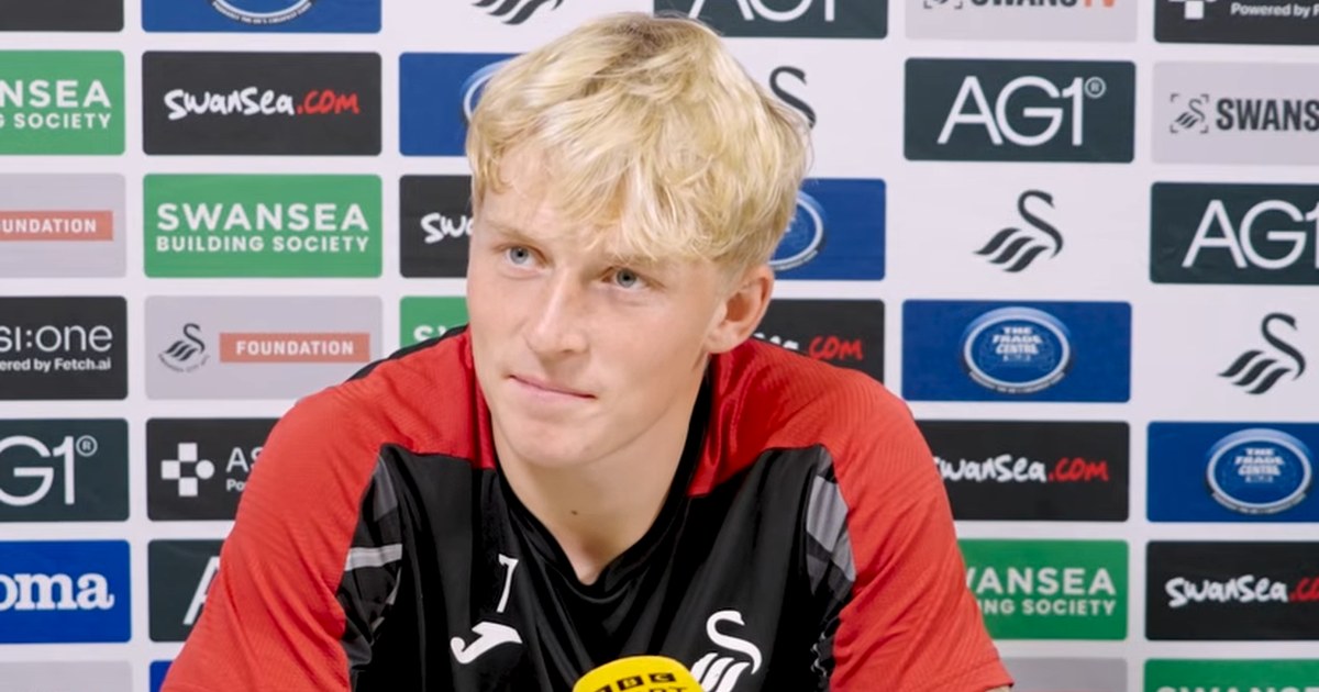 Swansea City's Melker Widell at a press conference in front of a sponsor board, wearing a red and black training top