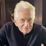 Michael Douglas, white-haired in a navy zip-neck jumper, leaning forward in an armchair in front of a cabinet of awards