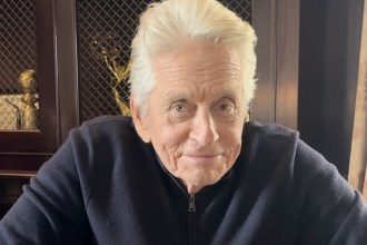 Michael Douglas, white-haired in a navy zip-neck jumper, leaning forward in an armchair in front of a cabinet of awards
