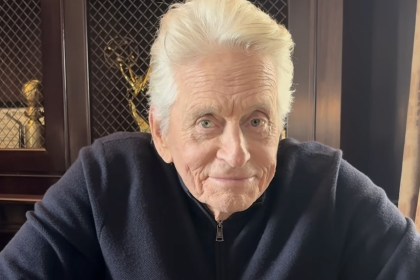 Michael Douglas, white-haired in a navy zip-neck jumper, leaning forward in an armchair in front of a cabinet of awards