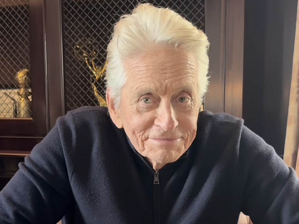 Michael Douglas, white-haired in a navy zip-neck jumper, leaning forward in an armchair in front of a cabinet of awards