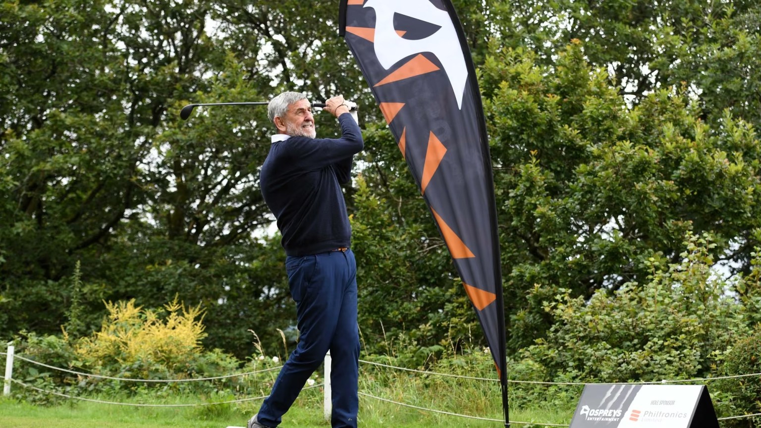 Mike James mid-swing with a golf club beside an Ospreys Entertainment flag at a golf day