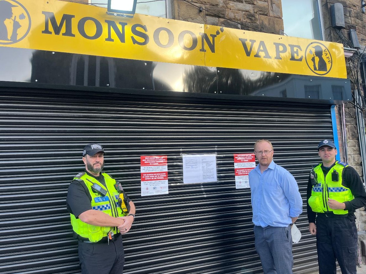 Swansea Council's lead trading standards officer Rhys Harries with two South Wales Police officers in front of the closed shutters of Monsoon Vape in Morriston, with court closure notices displayed
