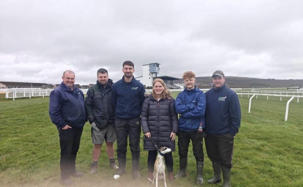 Morgan Hughes standing with five groundstaff colleagues and a whippet on the track at Ffos Las Racecourse, with the grandstand behind