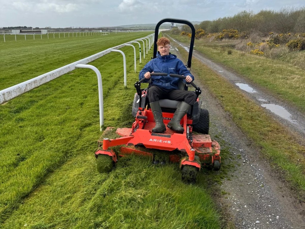 Morgan Hughes driving a red ride-on mower along the inside of the white running rail at Ffos Las Racecourse