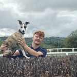 Morgan Hughes smiling behind a steeplechase fence at Ffos Las Racecourse, holding a brindle and white whippet sitting on top of the fence