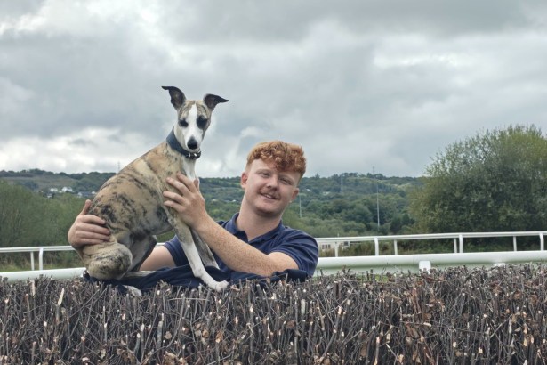 Morgan Hughes smiling behind a steeplechase fence at Ffos Las Racecourse, holding a brindle and white whippet sitting on top of the fence
