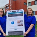 Two paediatric respiratory nurses in blue uniforms hold up a large Mould Matters poster outside the main entrance of Morriston Hospital