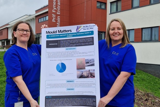 Two paediatric respiratory nurses in blue uniforms hold up a large Mould Matters poster outside the main entrance of Morriston Hospital