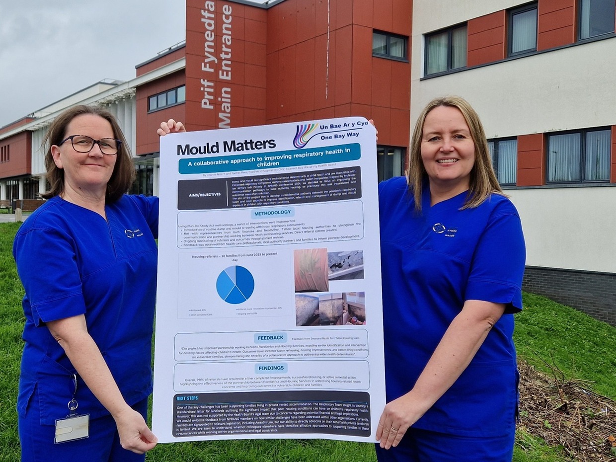 Two paediatric respiratory nurses in blue uniforms hold up a large Mould Matters poster outside the main entrance of Morriston Hospital