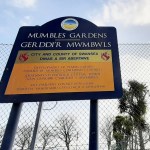 Blue and yellow Swansea Council sign reading Mumbles Gardens, Gerddi'r Mwmbwls, on the chain-link fence of the old tennis courts, saying the courts were funded by Mumbles Community Council