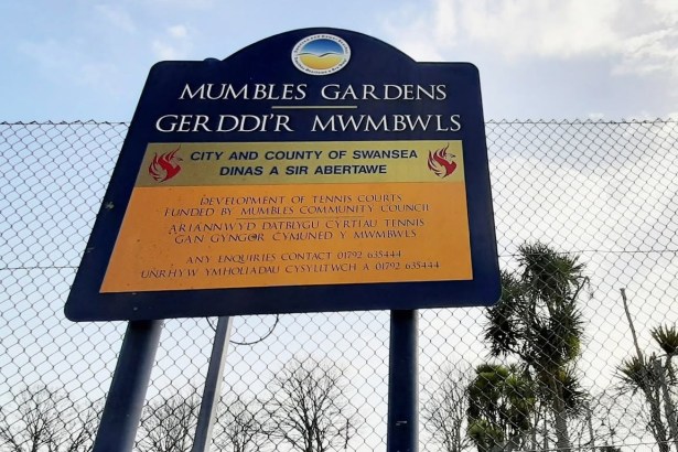 Blue and yellow Swansea Council sign reading Mumbles Gardens, Gerddi'r Mwmbwls, on the chain-link fence of the old tennis courts, saying the courts were funded by Mumbles Community Council