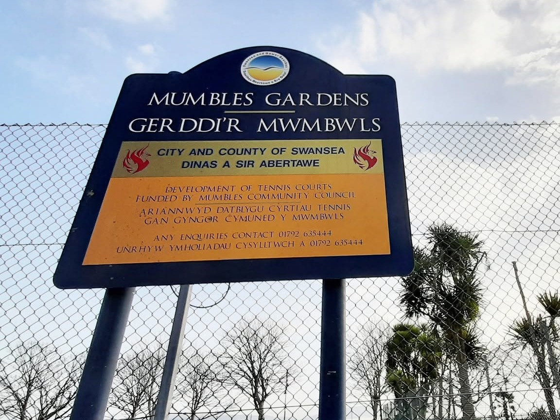 Blue and yellow Swansea Council sign reading Mumbles Gardens, Gerddi'r Mwmbwls, on the chain-link fence of the old tennis courts, saying the courts were funded by Mumbles Community Council