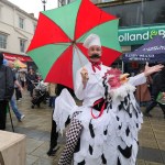 A performer in a chef's hat and false moustache holding a red and green umbrella, in a costume that makes him look like he is riding a giant chicken, on a wet Neath street with stalls behind