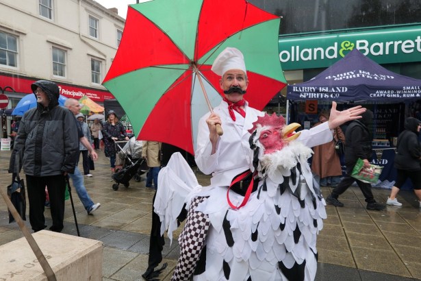 A performer in a chef's hat and false moustache holding a red and green umbrella, in a costume that makes him look like he is riding a giant chicken, on a wet Neath street with stalls behind