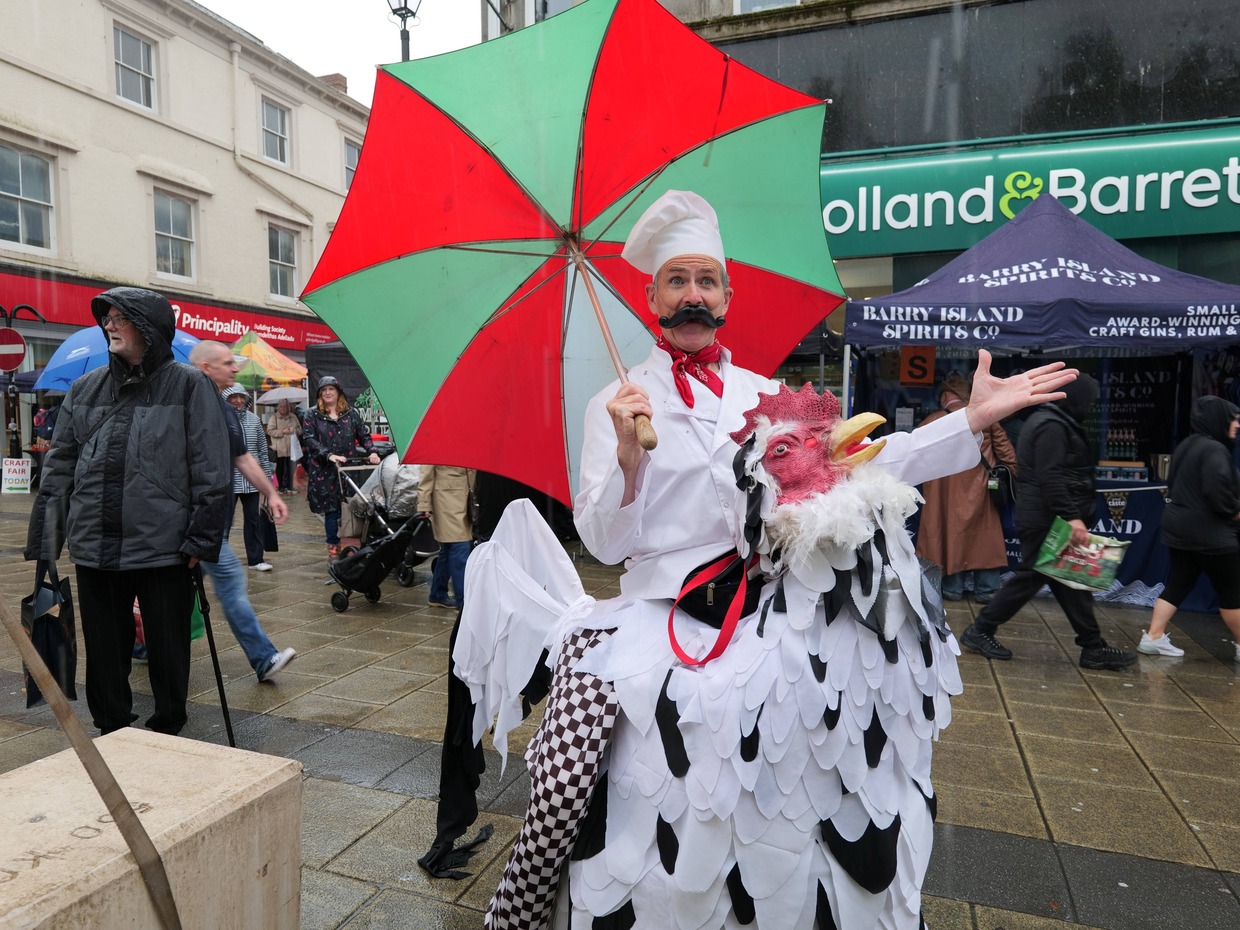 A performer in a chef's hat and false moustache holding a red and green umbrella, in a costume that makes him look like he is riding a giant chicken, on a wet Neath street with stalls behind