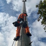 An engineer in orange overalls and a white helmet, harnessed with ropes, climbs a tall grey railway radio mast against a blue sky, with the weather station visible at the top