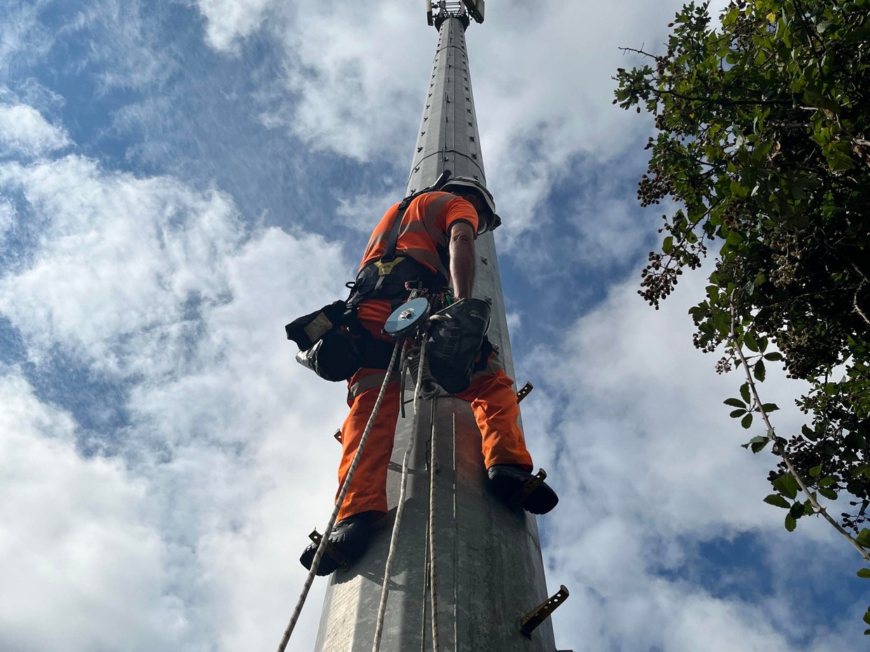 An engineer in orange overalls and a white helmet, harnessed with ropes, climbs a tall grey railway radio mast against a blue sky, with the weather station visible at the top