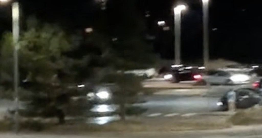 Blurred night-time video still of several cars with headlights on gathered in the North Dock car park, Llanelli, under street lights