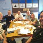 Seven members of the North Gate Swansea Pride in Place board smiling around a meeting table in a room with nautical decor