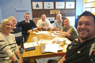 Seven members of the North Gate Swansea Pride in Place board smiling around a meeting table in a room with nautical decor