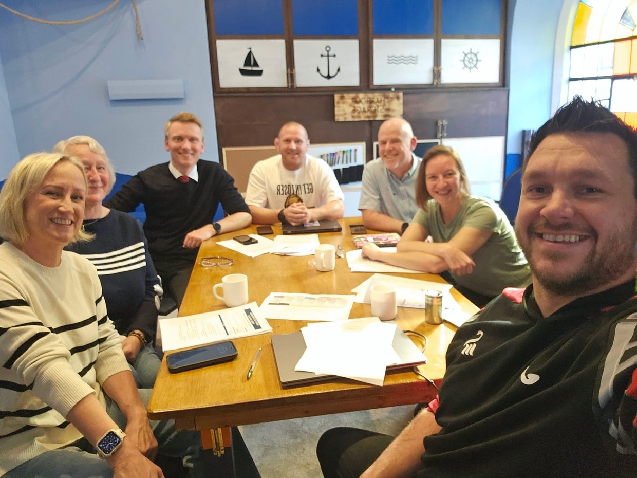 Seven members of the North Gate Swansea Pride in Place board smiling around a meeting table in a room with nautical decor