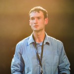 Oliver Jacob Davies in a blue shirt and lanyard, lit from behind in a warm haze, on stage in A Tidy Boy