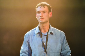 Oliver Jacob Davies in a blue shirt and lanyard, lit from behind in a warm haze, on stage in A Tidy Boy