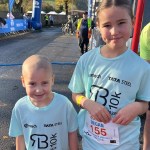 Olivia and Megan Finch in light blue Richard Burton 10K Mini Miler shirts with race numbers at the start line, the Tata Steel arch behind them