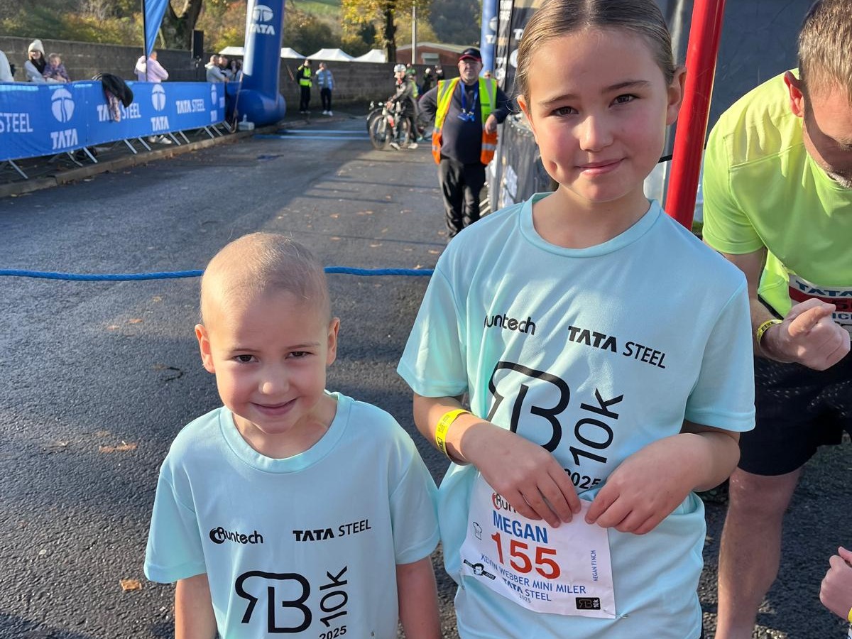 Olivia and Megan Finch in light blue Richard Burton 10K Mini Miler shirts with race numbers at the start line, the Tata Steel arch behind them